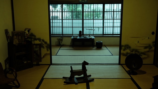 A traditional Japanese room with tatami mats covers the floor. An ink painting decorates one wall, while a low table and various decorative items fill the space. Natural light filters through a large window with wooden latticework, casting a serene glow.