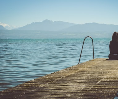 a woman sits on the end of a dock during daytime staring across a lake