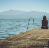 a woman sits on the end of a dock during daytime staring across a lake