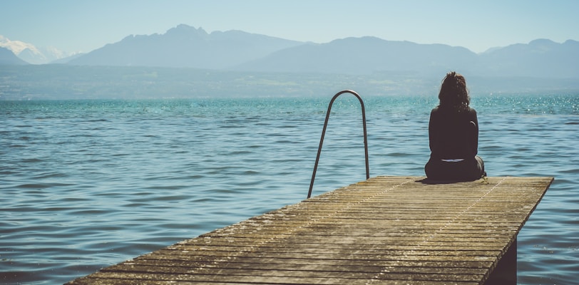 a woman sits on the end of a dock during daytime staring across a lake