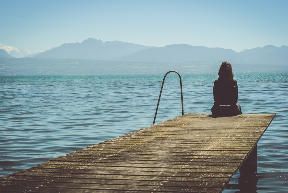 Eine Frau sitzt tagsüber am Ende eines Stegs und starrt über einen See