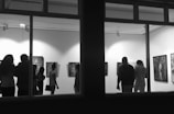 A group of people are observing artworks displayed on the walls of a gallery. The room is well-lit from the ceiling, and various framed pieces of art are visible, including portraits and abstract compositions. The scene is viewed from outside through large windows.