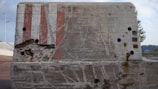 A large, weathered concrete barrier with visible wear and tear, including cracks and rust stains. It has holes drilled into it in various places and features faded red and white stripes on its surface. The background includes an overcast sky and distant greenery.