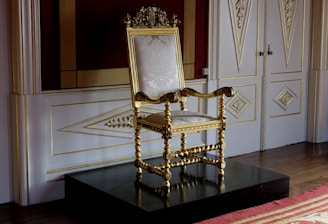 An ornate, gilded chair with elaborate carvings and twisted columns stands on a raised platform in a luxurious setting. The chair features intricate details and upholstery with a subtle floral pattern, surrounded by finely decorated wood paneling and geometric designs on the walls.