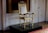 An ornate, gilded chair with elaborate carvings and twisted columns stands on a raised platform in a luxurious setting. The chair features intricate details and upholstery with a subtle floral pattern, surrounded by finely decorated wood paneling and geometric designs on the walls.
