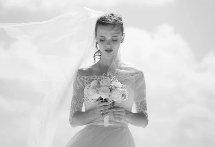 A black-and-white editorial portrait of a bride at her fitting, veil and bouquet in soft light