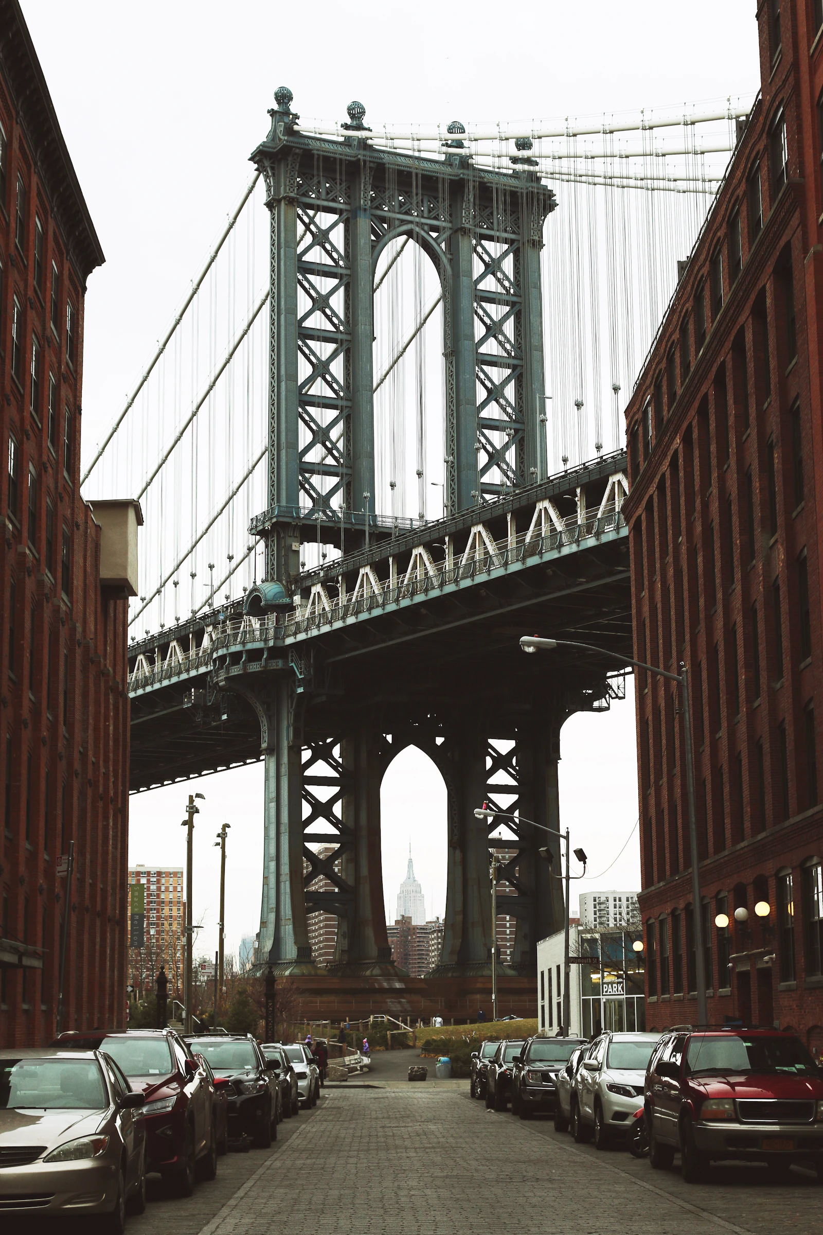 Travel photograph: The Manhattan Bridge framed by red-brick DUMBO warehouses, with a distant tower beneath its arch.