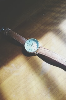 A classic wristwatch with a brown leather strap is placed on a wooden surface. The watch face is pale with black hour markers and hands, and a small logo near the top. Sunlight casts a soft shadow, creating a warm ambiance.