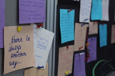 A series of colorful paper notes pinned to a fabric-covered board. Each note contains handwritten messages, with various colors like purple, blue, and beige paper. The notes are attached using push pins of different colors, including red, yellow, and blue. The visible texts promote positivity and inspiration.