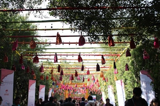 A decorated outdoor pathway features a colorful canopy of hanging fabric pouches and strings, with lush green trees lining the path. Banners and signs with logos are visible on either side, and people are walking below, possibly attending an event.
