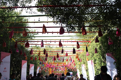 A decorated outdoor pathway features a colorful canopy of hanging fabric pouches and strings, with lush green trees lining the path. Banners and signs with logos are visible on either side, and people are walking below, possibly attending an event.