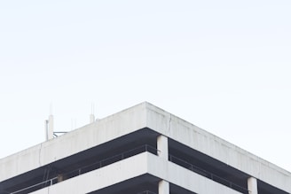 A minimalist view of a concrete building structure against a clear, pale sky. The building features simple geometric lines and visible structural elements like beams and columns. The surface of the concrete appears slightly weathered, emphasizing the industrial nature of the architecture.