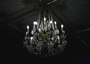 A luxurious crystal chandelier with multiple glowing bulbs hanging from an ornate ceiling. The chandelier features intricate designs with gleaming crystals and golden decorations, casting a soft light in a dimly lit room.