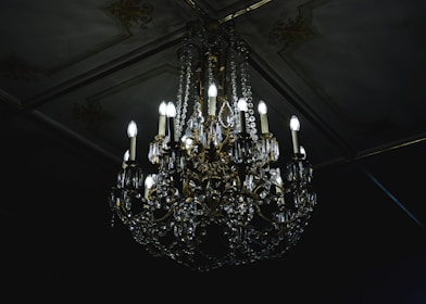 A luxurious crystal chandelier with multiple glowing bulbs hanging from an ornate ceiling. The chandelier features intricate designs with gleaming crystals and golden decorations, casting a soft light in a dimly lit room.