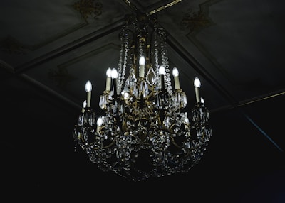 A luxurious crystal chandelier with multiple glowing bulbs hanging from an ornate ceiling. The chandelier features intricate designs with gleaming crystals and golden decorations, casting a soft light in a dimly lit room.