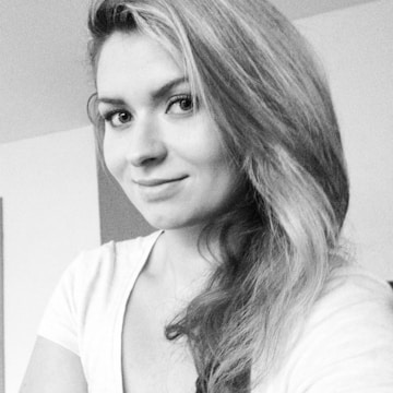 A grayscale portrait of a person with long hair, slightly smiling and looking towards the camera. The background appears to be indoors with simple decor.