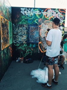 A person in casual clothing is creating art on a wall with vibrant graffiti that includes abstract and elaborate designs. Various spray paint patterns in bright colors cover the surfaces, and there are additional paintings leaned against the wall. Another person is partially visible in the background, and art supplies, including spray cans, are on the ground.