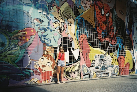A vibrant and colorful mural depicting various comic book characters and animated figures on a tiled wall. A person wearing sunglasses, a red tank top, denim shorts, and a crossbody bag stands in front of the mural, creating a contrast between the real and animated worlds.