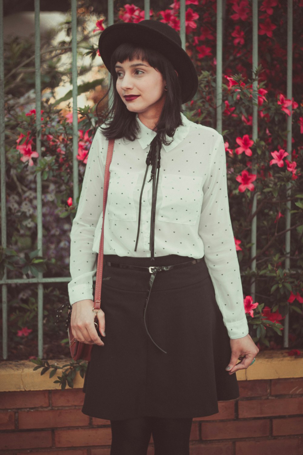 woman in white dress shirt and black skirt standing near fence with red flowers