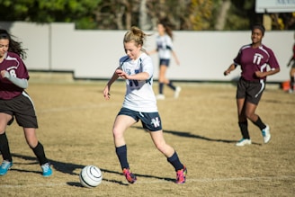 A young soccer player in a white jersey and blue shorts is dribbling a soccer ball on a grassy field, surrounded by opposing players in maroon jerseys. The scene suggests active gameplay with focused expressions and dynamic movement. The background includes a blurred fence and trees.