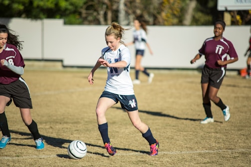 A young soccer player in a white jersey and blue shorts is dribbling a soccer ball on a grassy field, surrounded by opposing players in maroon jerseys. The scene suggests active gameplay with focused expressions and dynamic movement. The background includes a blurred fence and trees.