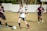 A young soccer player in a white jersey and blue shorts is dribbling a soccer ball on a grassy field, surrounded by opposing players in maroon jerseys. The scene suggests active gameplay with focused expressions and dynamic movement. The background includes a blurred fence and trees.