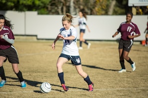 A young soccer player in a white jersey and blue shorts is dribbling a soccer ball on a grassy field, surrounded by opposing players in maroon jerseys. The scene suggests active gameplay with focused expressions and dynamic movement. The background includes a blurred fence and trees.