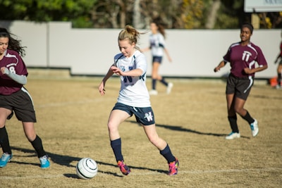 A young soccer player in a white jersey and blue shorts is dribbling a soccer ball on a grassy field, surrounded by opposing players in maroon jerseys. The scene suggests active gameplay with focused expressions and dynamic movement. The background includes a blurred fence and trees.