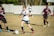 A young soccer player in a white jersey and blue shorts is dribbling a soccer ball on a grassy field, surrounded by opposing players in maroon jerseys. The scene suggests active gameplay with focused expressions and dynamic movement. The background includes a blurred fence and trees.