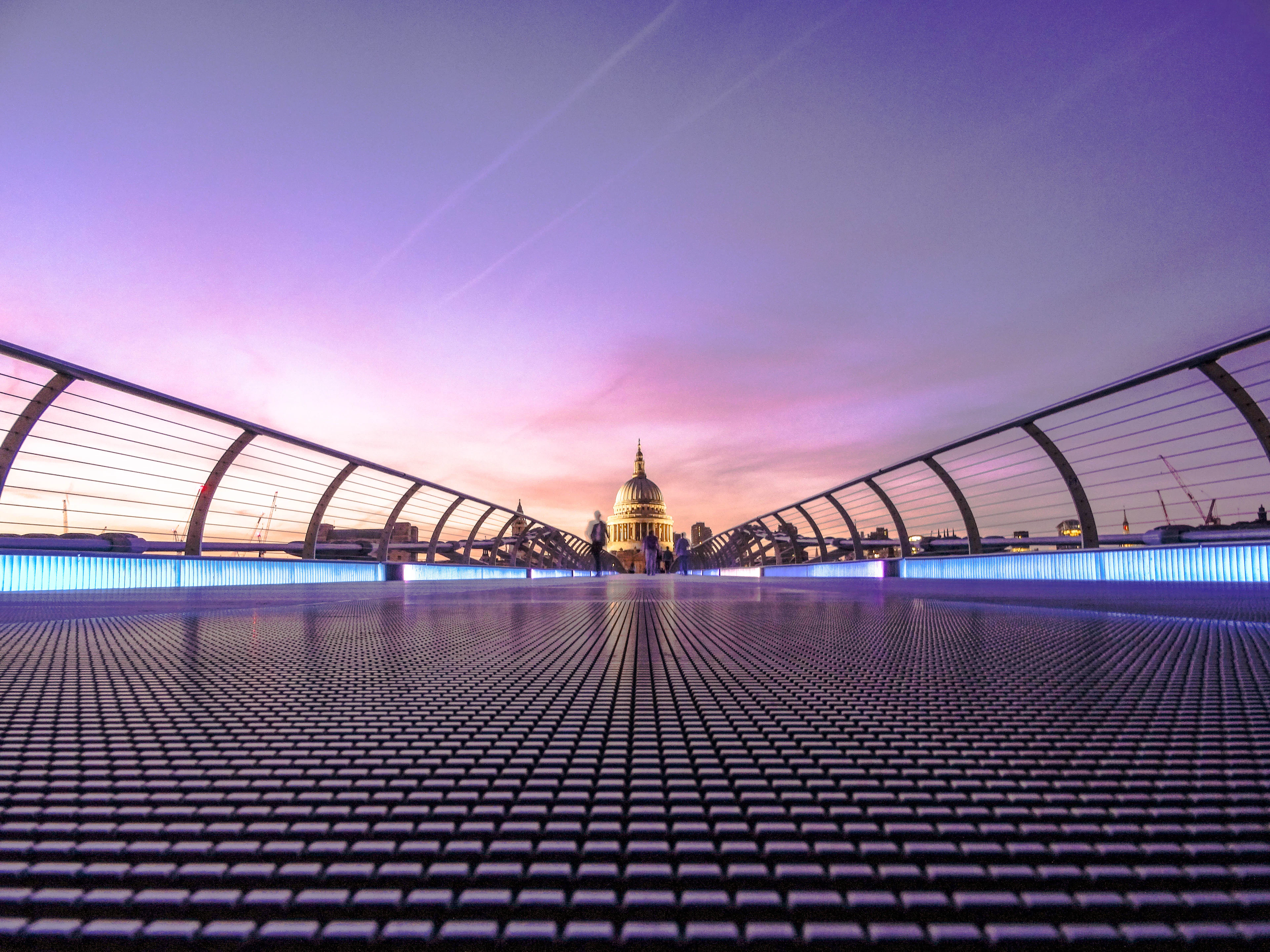 The Millennium Bridge's steel deck at dusk leading toward the floodlit dome of St Paul's Cathedral under a violet London sky