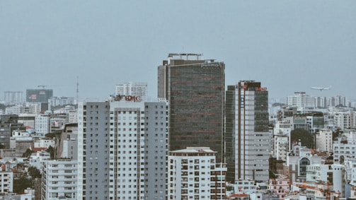 A dense cityscape featuring multiple high-rise buildings, including a prominent dark building with the 'Pullman' signage and a white building with 'BMC' sign. The skyline indicates numerous residential and commercial structures, with a few planes in the sky indicating an urban area near an airport.