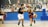 Two young basketball players are in action on an indoor court. One player in a black jersey is attempting to block another player in a blue and red jersey who is dribbling a basketball. Other players and a basketball hoop can be seen in the background. The court is surrounded by a blue wall with sports-themed murals above it.