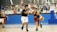 Two young basketball players are in action on an indoor court. One player in a black jersey is attempting to block another player in a blue and red jersey who is dribbling a basketball. Other players and a basketball hoop can be seen in the background. The court is surrounded by a blue wall with sports-themed murals above it.