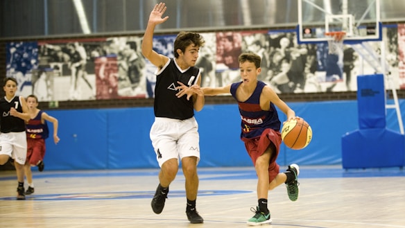 Two young basketball players are in action on an indoor court. One player in a black jersey is attempting to block another player in a blue and red jersey who is dribbling a basketball. Other players and a basketball hoop can be seen in the background. The court is surrounded by a blue wall with sports-themed murals above it.