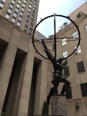A bronze statue depicting a muscular man holding a large celestial sphere on his shoulders, set against the backdrop of tall, urban architecture. The buildings have a grid of windows and a neutral stone color.