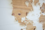 A wooden world map with details of North and Central America, featuring labeled countries such as Canada, the United States, and Mexico. Several colorful pins are placed on the map, marking specific locations. The map displays a vintage aesthetic with a beige and brown color palette.