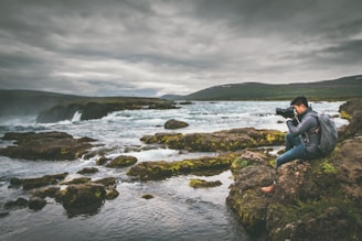 A serene scene of a person sitting on rocky terrain, focused on photography against the backdrop of a flowing river and cloudy sky. The landscape is lush with patches of green moss and surrounded by the expansive river flowing over rocks.