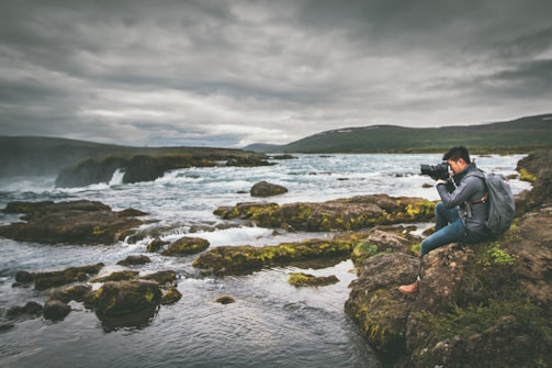 A serene scene of a person sitting on rocky terrain, focused on photography against the backdrop of a flowing river and cloudy sky. The landscape is lush with patches of green moss and surrounded by the expansive river flowing over rocks.