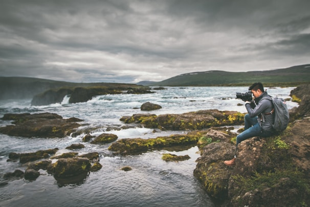 A serene scene of a person sitting on rocky terrain, focused on photography against the backdrop of a flowing river and cloudy sky. The landscape is lush with patches of green moss and surrounded by the expansive river flowing over rocks.
