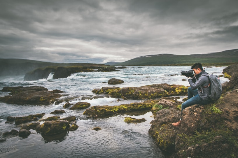 A serene scene of a person sitting on rocky terrain, focused on photography against the backdrop of a flowing river and cloudy sky. The landscape is lush with patches of green moss and surrounded by the expansive river flowing over rocks.