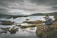 A serene scene of a person sitting on rocky terrain, focused on photography against the backdrop of a flowing river and cloudy sky. The landscape is lush with patches of green moss and surrounded by the expansive river flowing over rocks.