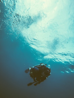 A scuba diver is submerged underwater with visible air bubbles rising towards the surface. The person is equipped with scuba gear including an oxygen tank. The water has a blue-green tint and light filters from above, creating a serene atmosphere.