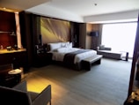 A luxurious hotel room with a large, neatly made bed adorned with white linens and grey accents. To the left side of the bed, there are two nightstands with lamps, and the wall behind the bed features a modern decorative design. On the right side, there is a sitting area with a long sofa and side table in front of a large window, allowing plenty of natural light to fill the room. A small dining table with two chairs is situated in the foreground on the left, next to a kitchenette area with shelves holding bottles and glassware.