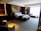 A luxurious hotel room with a large, neatly made bed adorned with white linens and grey accents. To the left side of the bed, there are two nightstands with lamps, and the wall behind the bed features a modern decorative design. On the right side, there is a sitting area with a long sofa and side table in front of a large window, allowing plenty of natural light to fill the room. A small dining table with two chairs is situated in the foreground on the left, next to a kitchenette area with shelves holding bottles and glassware.