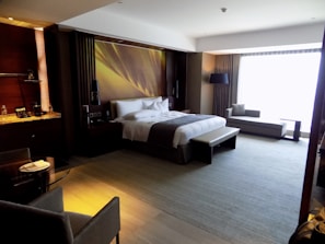 A luxurious hotel room with a large, neatly made bed adorned with white linens and grey accents. To the left side of the bed, there are two nightstands with lamps, and the wall behind the bed features a modern decorative design. On the right side, there is a sitting area with a long sofa and side table in front of a large window, allowing plenty of natural light to fill the room. A small dining table with two chairs is situated in the foreground on the left, next to a kitchenette area with shelves holding bottles and glassware.