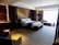 A luxurious hotel room with a large, neatly made bed adorned with white linens and grey accents. To the left side of the bed, there are two nightstands with lamps, and the wall behind the bed features a modern decorative design. On the right side, there is a sitting area with a long sofa and side table in front of a large window, allowing plenty of natural light to fill the room. A small dining table with two chairs is situated in the foreground on the left, next to a kitchenette area with shelves holding bottles and glassware.