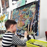A young child is seated at an easel, working on a colorful painting using oil pastels. The painting features a vibrant depiction of a chameleon with a dotted pattern in various colors such as orange, blue, and yellow, against a dark background. Nearby is a basket filled with art supplies, indicating an active engagement in the creative process. Behind, there are other finished paintings with sunflowers and still life, hanging on a wall.
