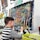 A young child is seated at an easel, working on a colorful painting using oil pastels. The painting features a vibrant depiction of a chameleon with a dotted pattern in various colors such as orange, blue, and yellow, against a dark background. Nearby is a basket filled with art supplies, indicating an active engagement in the creative process. Behind, there are other finished paintings with sunflowers and still life, hanging on a wall.
