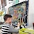 A young child is seated at an easel, working on a colorful painting using oil pastels. The painting features a vibrant depiction of a chameleon with a dotted pattern in various colors such as orange, blue, and yellow, against a dark background. Nearby is a basket filled with art supplies, indicating an active engagement in the creative process. Behind, there are other finished paintings with sunflowers and still life, hanging on a wall.