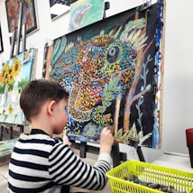 A young child is seated at an easel, working on a colorful painting using oil pastels. The painting features a vibrant depiction of a chameleon with a dotted pattern in various colors such as orange, blue, and yellow, against a dark background. Nearby is a basket filled with art supplies, indicating an active engagement in the creative process. Behind, there are other finished paintings with sunflowers and still life, hanging on a wall.
